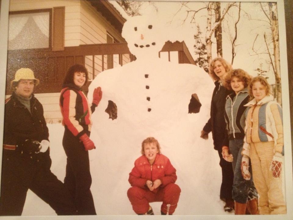 A young Lara in a red snowsuit, crouched in front of a snowman roughly the size of a garage, surrounded by family.