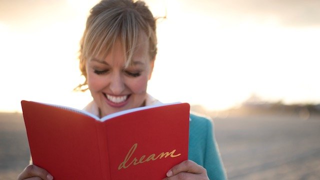 Lara on a windy beach at sunset, smiling down at a red journal with the word dream on the cover.