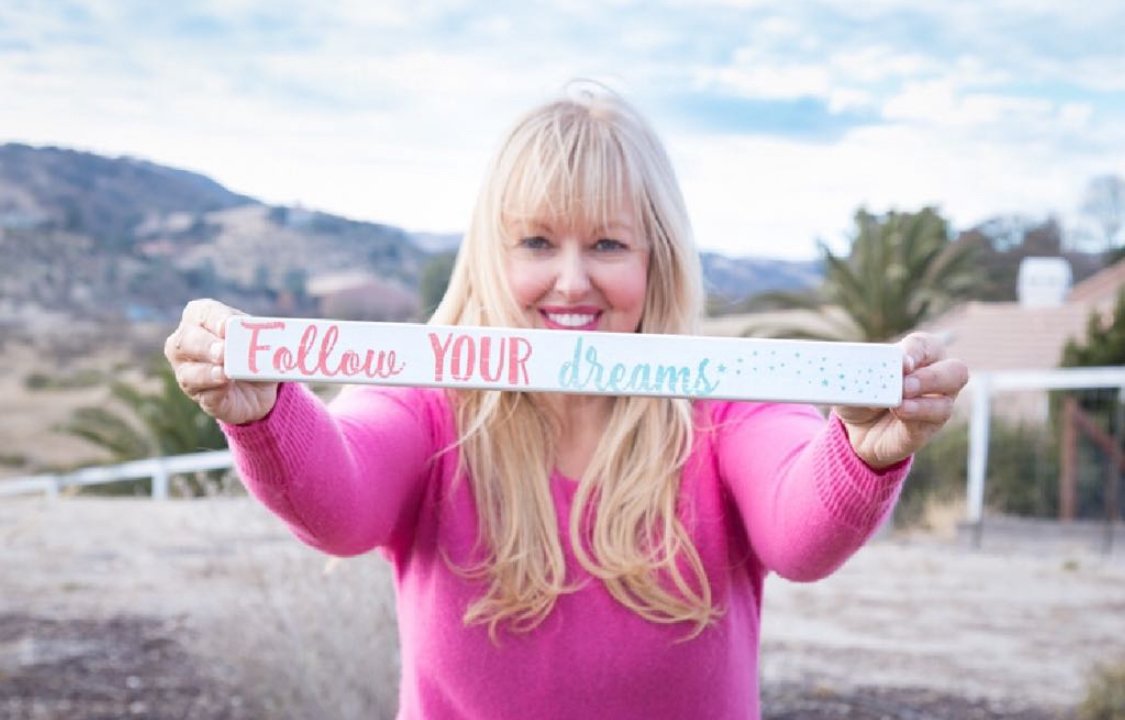 Lara in a pink sweater holding a hand-painted sign that reads 'Follow YOUR dreams,' hills behind her.