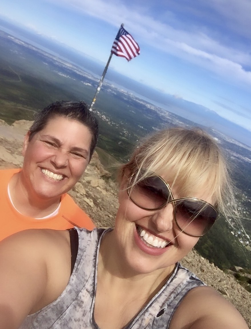 Lara and a grade-school friend laughing at the summit of a mountain, an American flag and wide valley behind them.